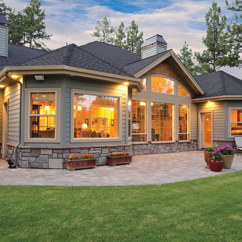 Exterior view of a custom home at dusk featuring large patio doors and windows, warm interior lighting, stone accents, and landscaped yard.