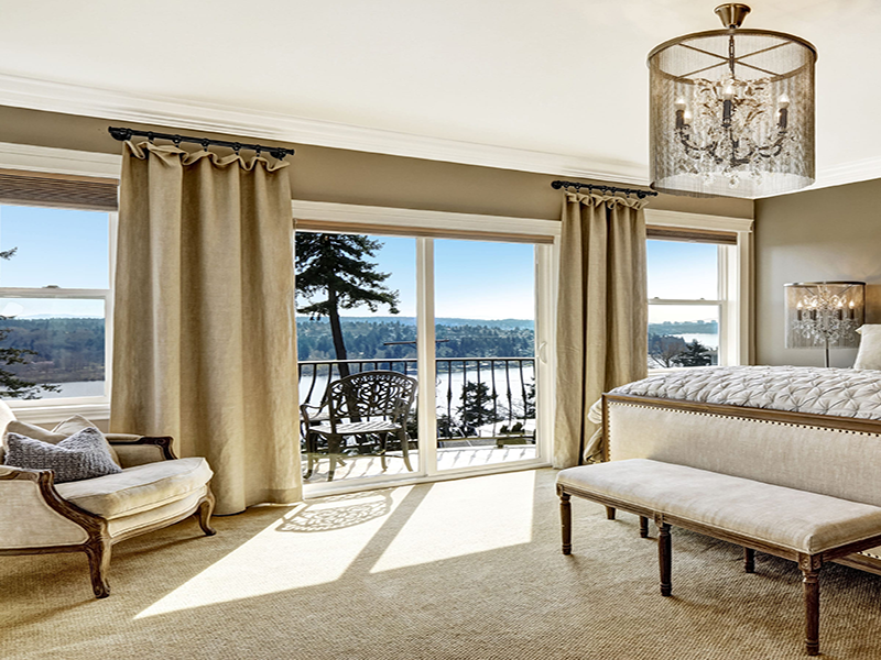 Elegant bedroom featuring large sliding glass patio doors with curtains, opening to a private balcony with scenic lake and tree views.